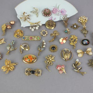 May include: A collection of assorted vintage-style metal brooches in various shapes including flowers, bows, and abstract designs. Many pieces feature gold-tone metal, faux pearls, colorful rhinestones, and enamel accents, arranged on a white fan-shaped base.