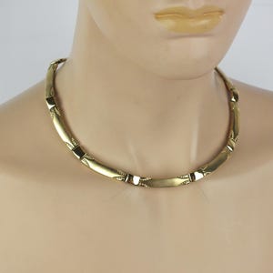 K&ouml;nnte beinhalten: Eine goldfarbene Halskette mit einer Reihe von verbundenen, gebogenen rechteckigen Gliedern. Die Halskette wird um den Hals getragen und hat ein schlichtes, elegantes Design. Die Glieder sind durch kleinere, dekorative Elemente verbunden.