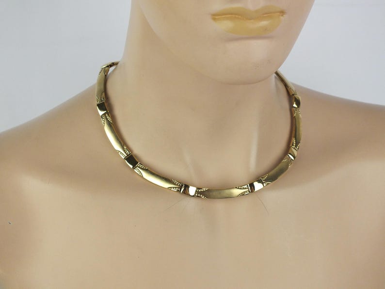 K&ouml;nnte beinhalten: Eine goldfarbene Halskette mit einer Reihe von verbundenen, gebogenen rechteckigen Gliedern. Die Halskette wird um den Hals getragen und hat ein schlichtes, elegantes Design. Die Glieder sind durch kleinere, dekorative Elemente verbunden.