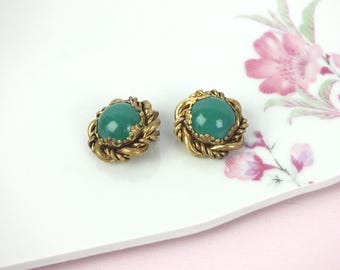 May include: A pair of round earrings featuring a smooth, domed green cabochon stone set in the center. The stone is surrounded by a textured, twisted gold-tone metal border with a scalloped edge detail that frames the central green element.