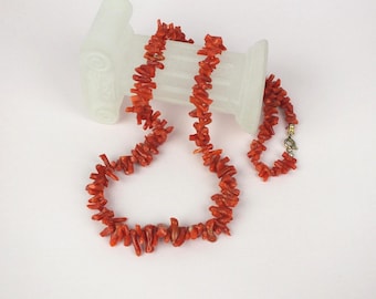 May include: A vibrant red coral necklace and matching bracelet. The necklace and bracelet are composed of irregular, natural coral pieces. The necklace is draped on a white surface, with a decorative white object in the background. The bracelet is fastened with a gold-colored clasp.