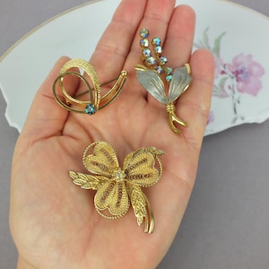 May include: Three gold-tone metal brooches. One brooch features a swirling design with a small blue stone. Another is a floral-shaped piece with silver-toned leaves and iridescent blue crystals. The third is a filigree brooch with a central stone.