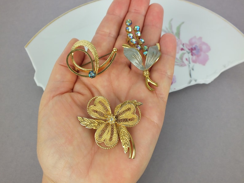 May include: Three gold-tone metal brooches. One brooch features a swirling design with a small blue stone. Another is a floral-shaped piece with silver-toned leaves and iridescent blue crystals. The third is a filigree brooch with a central stone.