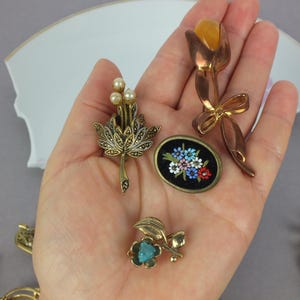 May include: A collection of four distinct brooches: one featuring a gold-tone tulip with an amber-colored stone, one with three small pearls and marcasite accents, one oval black brooch with floral details, and one small gold-tone flower with a blue stone.