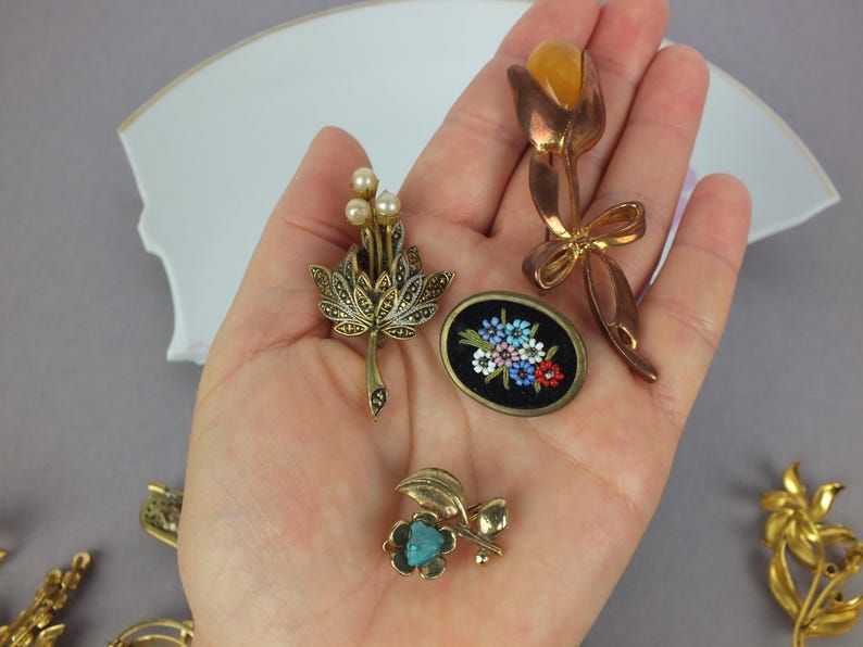 May include: A collection of four distinct brooches: one featuring a gold-tone tulip with an amber-colored stone, one with three small pearls and marcasite accents, one oval black brooch with floral details, and one small gold-tone flower with a blue stone.