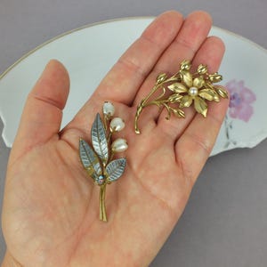 May include: Two floral brooches made of gold-toned metal. One brooch features three white pearl-like beads on stems with textured silver-toned leaves. The second brooch is a gold-toned flower with a central pearl-like bead and surrounding buds.