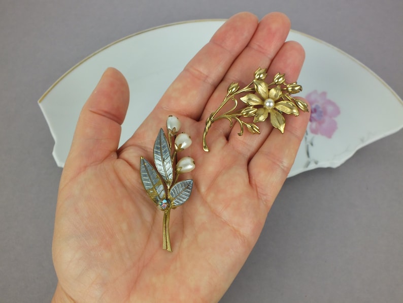 May include: Two floral brooches made of gold-toned metal. One brooch features three white pearl-like beads on stems with textured silver-toned leaves. The second brooch is a gold-toned flower with a central pearl-like bead and surrounding buds.