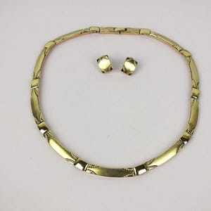 K&ouml;nnte beinhalten: Ein goldfarbenes Schmuckset mit Halskette und passenden Ohrringen. Die Halskette ist im Choker-Stil mit rechteckigen Gliedern und dekorativen Details gehalten. Die Ohrringe sind kleine, quadratische Ohrstecker. Das Set wird auf einer wei&szlig;en Oberfl&auml;che pr&auml;sentiert.