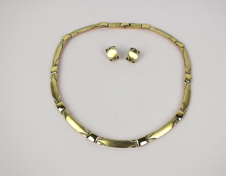 K&ouml;nnte beinhalten: Ein goldfarbenes Schmuckset mit Halskette und passenden Ohrringen. Die Halskette ist im Choker-Stil mit rechteckigen Gliedern und dekorativen Details gehalten. Die Ohrringe sind kleine, quadratische Ohrstecker. Das Set wird auf einer wei&szlig;en Oberfl&auml;che pr&auml;sentiert.
