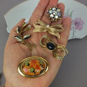 May include: A collection of five gold-tone brooches including a round cluster of iridescent rhinestones, a textured bow with a central pearl, a floral spray with white enamel, a bow with a black oval stone and clear rhinestones, and an oval floral pin.