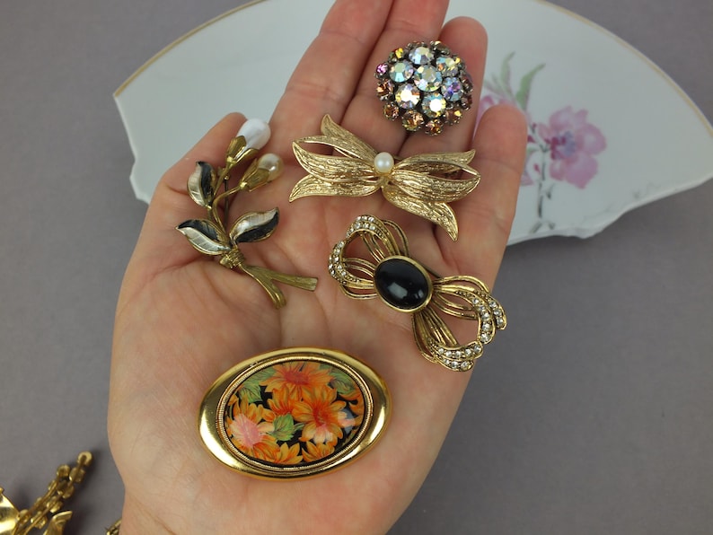 May include: A collection of five gold-tone brooches including a round cluster of iridescent rhinestones, a textured bow with a central pearl, a floral spray with white enamel, a bow with a black oval stone and clear rhinestones, and an oval floral pin.