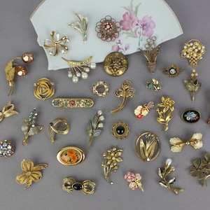 May include: A collection of assorted vintage-style brooches in gold-tone metal, featuring floral, bow, and abstract designs with faux pearls, colorful rhinestones, and enamel accents, arranged around a curved piece of white porcelain with floral art.