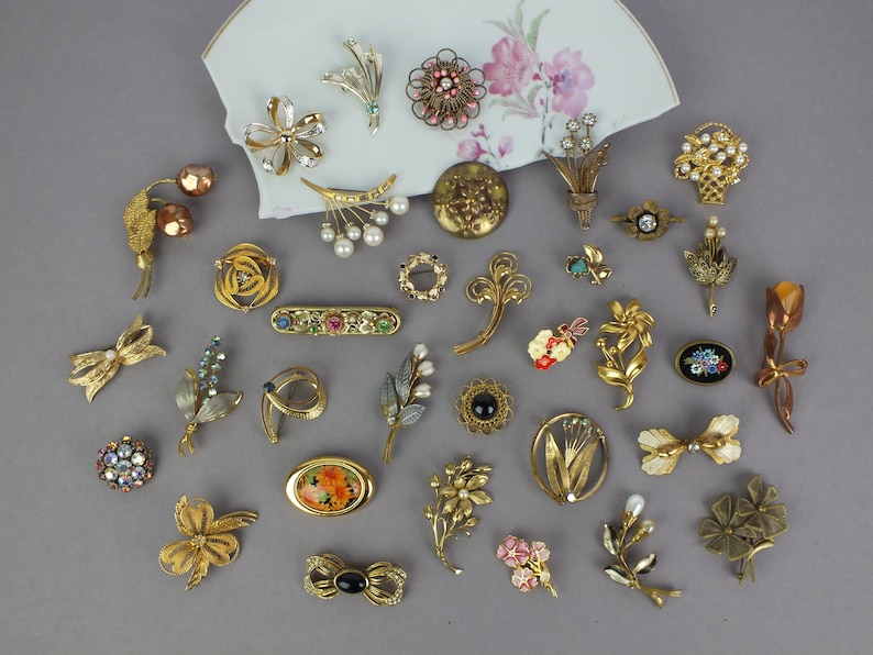 May include: A collection of assorted vintage-style brooches in gold-tone metal, featuring floral, bow, and abstract designs with faux pearls, colorful rhinestones, and enamel accents, arranged around a curved piece of white porcelain with floral art.