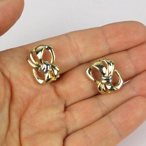 以下が含まれることがあります： 中央にシルバーカラーのアクセントを配した、結び目のようなループデザインのポリッシュ仕上げゴールドトーンイヤリング。滑らかで光沢のある表面と、構造的で対称的な形状が特徴で、オープンで軽やかな印象を与えます。