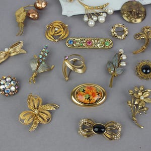 May include: A collection of assorted gold-tone metal brooches featuring various designs including floral motifs, bows, and geometric shapes. The pieces are embellished with colorful rhinestones, faux pearls, black cabochons, and painted enamel floral patterns.
