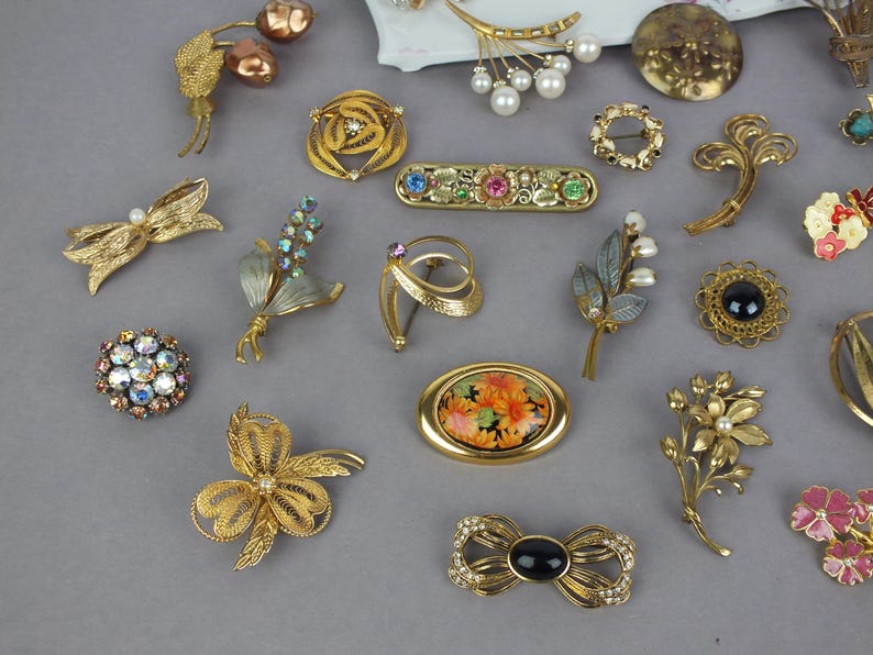 May include: A collection of assorted gold-tone metal brooches featuring various designs including floral motifs, bows, and geometric shapes. The pieces are embellished with colorful rhinestones, faux pearls, black cabochons, and painted enamel floral patterns.