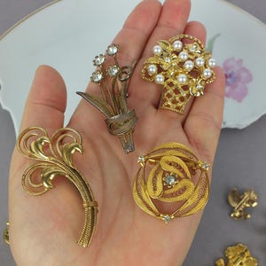 May include: A collection of four gold-tone metal brooches. One features a floral spray with clear rhinestones, another is a basket filled with white faux pearls, one is a swirling filigree design, and the last is a curved, textured leaf shape.