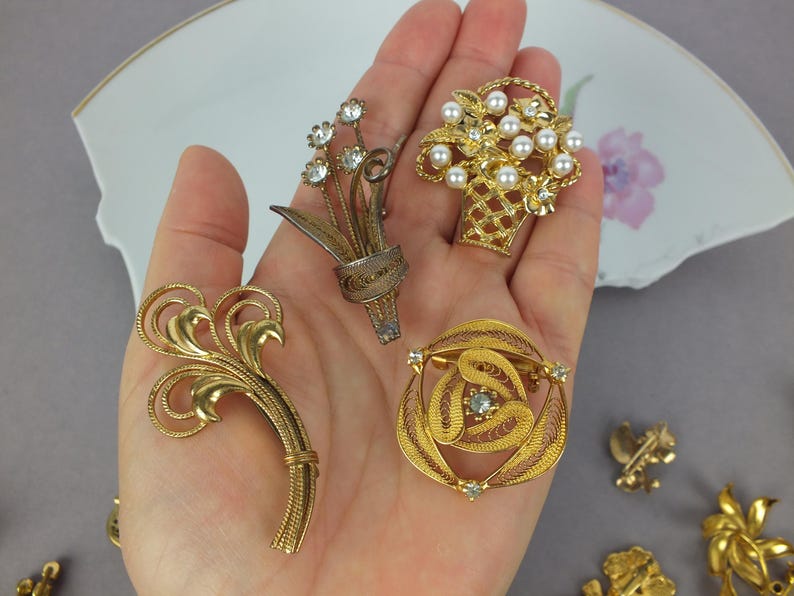 May include: A collection of four gold-tone metal brooches. One features a floral spray with clear rhinestones, another is a basket filled with white faux pearls, one is a swirling filigree design, and the last is a curved, textured leaf shape.