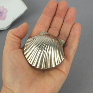 Könnte beinhalten: Eine silberfarbene Schmuckdose aus Metall in Form einer Jakobsmuschel. Das Objekt verfügt über ein Klappdesign mit einer gerippten, fächerartigen Textur auf der Außenfläche, die das Licht auf ihrer geschwungenen, metallischen Form reflektiert.