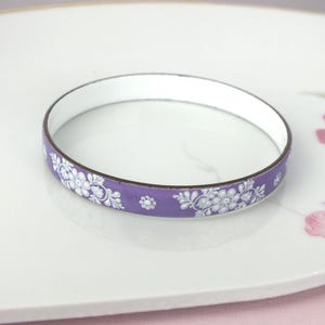 Könnte beinhalten: Ein kreisförmiger Armreif mit einer leuchtend violetten Außenseite, die mit einem zarten weißen Blumenmuster verziert ist. Die Innenseite des Armreifs ist einfarbig weiß und die Kanten sind mit einem dünnen, dunklen Metallrand versehen.