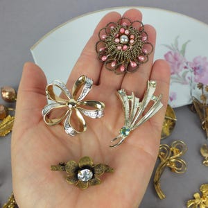 May include: A collection of four metal brooches featuring a round pink and gold beaded pin, a gold and silver ribbon-shaped pin, a silver-toned spray pin with small crystals, and a small gold-toned flower pin with a central clear stone.