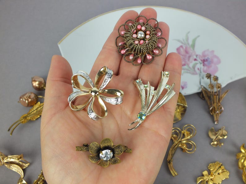 May include: A collection of four metal brooches featuring a round pink and gold beaded pin, a gold and silver ribbon-shaped pin, a silver-toned spray pin with small crystals, and a small gold-toned flower pin with a central clear stone.