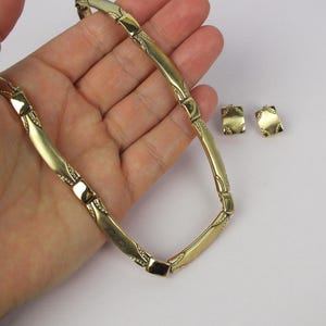 K&ouml;nnte beinhalten: Eine goldfarbene Halskette und passende quadratische Ohrringe. Die Halskette hat eine Reihe von gebogenen, rechteckigen Gliedern mit dekorativen Details. Die Ohrringe haben ein &auml;hnliches Design. Der Schmuck wird auf hellem Hintergrund pr&auml;sentiert.