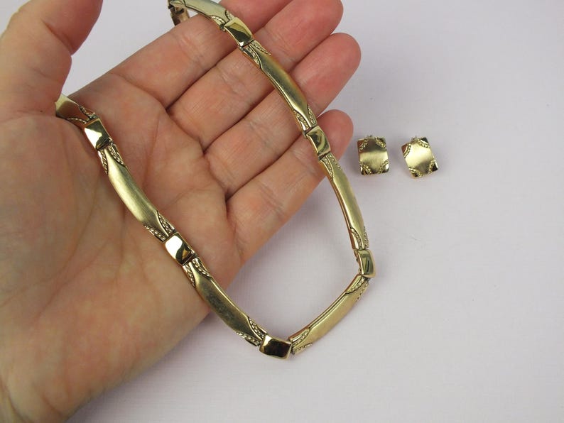 K&ouml;nnte beinhalten: Eine goldfarbene Halskette und passende quadratische Ohrringe. Die Halskette hat eine Reihe von gebogenen, rechteckigen Gliedern mit dekorativen Details. Die Ohrringe haben ein &auml;hnliches Design. Der Schmuck wird auf hellem Hintergrund pr&auml;sentiert.