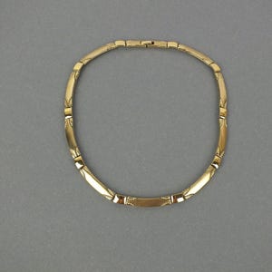 K&ouml;nnte beinhalten: Eine goldfarbene Halskette mit geometrischem Design. Die Halskette hat rechteckige und gebogene Segmente, die durch kleine Glieder verbunden sind. Die Segmente haben ge&auml;tzte dekorative Muster. Die Halskette wird auf grauem Hintergrund pr&auml;sentiert.