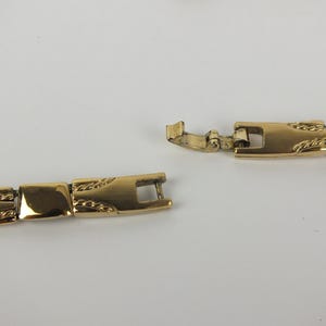K&ouml;nnte beinhalten: Eine goldfarbene Halskette mit rechteckigem Gliederdesign. Der Verschluss ist ge&ouml;ffnet und zeigt den Verbindungsmechanismus. Die Glieder haben ein strukturiertes Muster. Die Halskette wird vor wei&szlig;em Hintergrund pr&auml;sentiert.