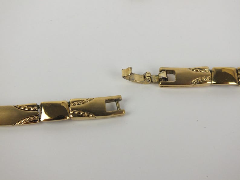 K&ouml;nnte beinhalten: Eine goldfarbene Halskette mit rechteckigem Gliederdesign. Der Verschluss ist ge&ouml;ffnet und zeigt den Verbindungsmechanismus. Die Glieder haben ein strukturiertes Muster. Die Halskette wird vor wei&szlig;em Hintergrund pr&auml;sentiert.