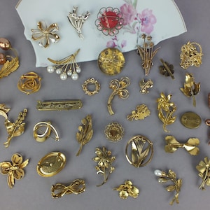 May include: A collection of various gold-toned metal brooches and pins featuring floral, leaf, and abstract designs. Some pieces include faux pearls, clear crystals, and textured finishes. The items are arranged on a white ceramic fragment.