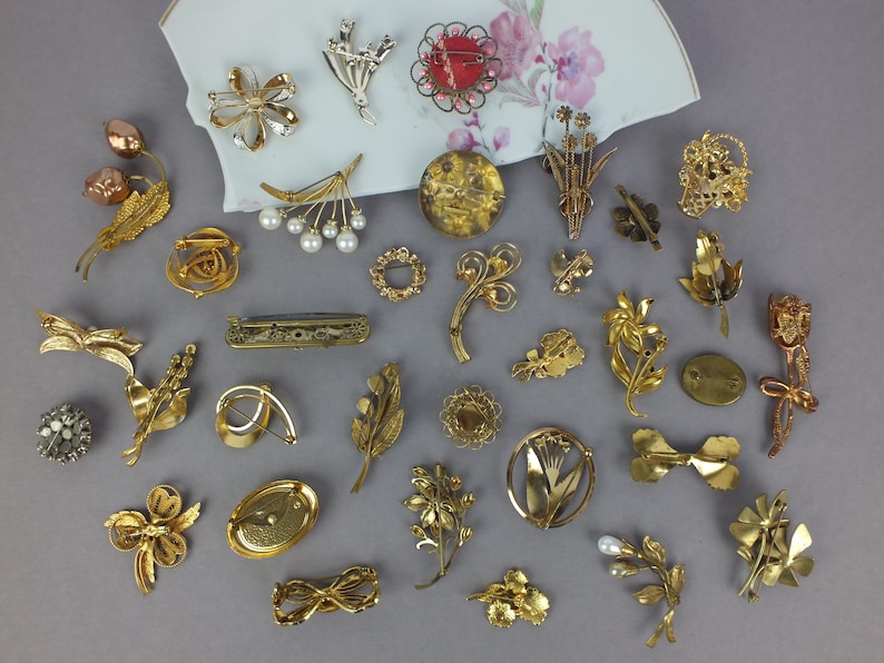 May include: A collection of various gold-toned metal brooches and pins featuring floral, leaf, and abstract designs. Some pieces include faux pearls, clear crystals, and textured finishes. The items are arranged on a white ceramic fragment.