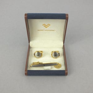 May include: A Mario Valentino set comprising cufflinks, a tie clip, and a small coin, displayed in a blue and brown box. The cufflinks and tie clip feature a gold-tone finish with a silver-tone centre. The box is lined with cream-coloured fabric.