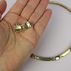 K&ouml;nnte beinhalten: Ein goldfarbenes Schmuckset mit einer Halskette und passenden Ohrringen. Die Halskette besteht aus rechteckigen Gliedern mit dekorativen Details. Die Ohrringe sind quadratisch geformt und weisen &auml;hnliche Verzierungen auf. Das Set wird auf hellem Hintergrund gezeigt.