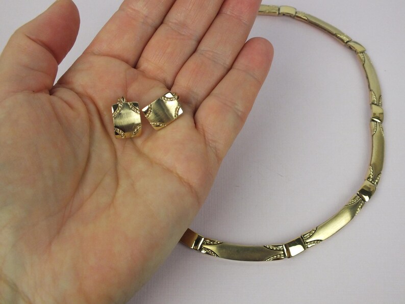 K&ouml;nnte beinhalten: Ein goldfarbenes Schmuckset mit einer Halskette und passenden Ohrringen. Die Halskette besteht aus rechteckigen Gliedern mit dekorativen Details. Die Ohrringe sind quadratisch geformt und weisen &auml;hnliche Verzierungen auf. Das Set wird auf hellem Hintergrund gezeigt.