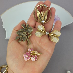 May include: A collection of four metal brooches including a circular floral design with crystals, a textured bow-shaped piece, a multi-petaled flower brooch with pink enamel, and a dark metallic floral pin with textured leaves and stems.
