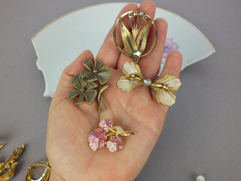 May include: A collection of four metal brooches including a circular floral design with crystals, a textured bow-shaped piece, a multi-petaled flower brooch with pink enamel, and a dark metallic floral pin with textured leaves and stems.
