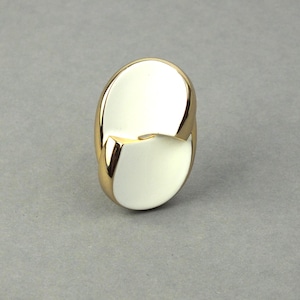 May include: A gold ring with a distinctive design. The ring has two overlapping, oval sections in a creamy white, framed by polished gold metal. The ring is set against a grey background.