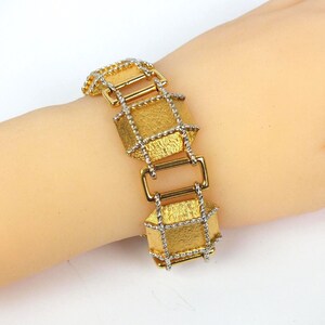 May include: A gold-coloured bracelet with a textured, geometric design. The bracelet features rectangular links with a woven silver-coloured trim and gold-coloured connecting bars. The bracelet is displayed on a light-toned surface.