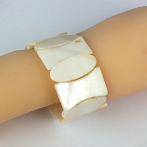 Könnte beinhalten: Cremefarbenes Armband mit rechteckigen und ovalen Segmenten. Das Armband aus glänzendem Material wird am Handgelenk getragen. Die Segmente sind durch kleine, flexible Glieder verbunden. Es ist ein Schmuckstück.