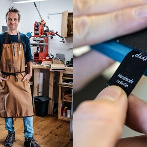 May include: A man wearing a brown leather work apron stands in a workshop. He is looking at the camera. The apron has multiple pockets and a tool loop. The workshop has a large red printing press in the background.