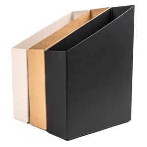 May include: Three cardboard magazine holders, one white, one brown, and one black, standing side by side against a white background.