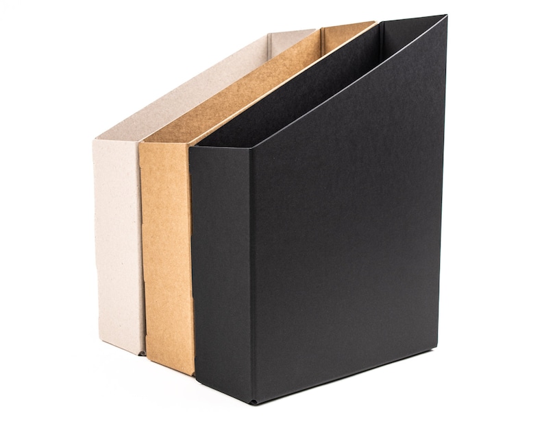 May include: Three cardboard magazine holders, one white, one brown, and one black, standing side by side against a white background.