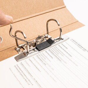 May include: A brown cardboard binder with a metal mechanism for holding papers. The binder is open and the metal mechanism is visible. The text on the page reads "KONZERN-GEWINN-UND-VERLUSTRECHNUN" and "F&uuml;r das Gesch&auml;ftsjahr 2007".