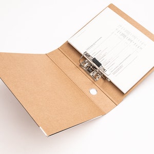 May include: A brown cardboard file folder with a metal ring binder and a white sheet of paper inside. The folder is open and the paper is visible.
