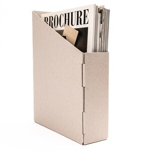 May include: A beige cardboard magazine holder with a stack of magazines inside. The top magazine has the word "BROCHURE" printed on the cover.
