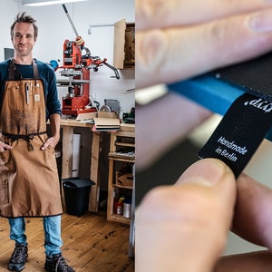 May include: A man wearing a brown leather work apron stands in a workshop. He is looking at the camera. The apron has multiple pockets and a tool loop. The workshop has a large red printing press in the background.