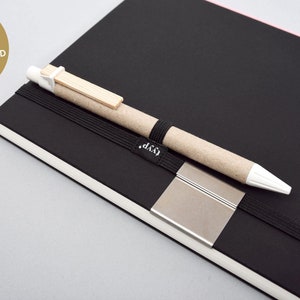 Può includere: Un taccuino nero con un portapenne e un elastico nero. Il taccuino ha una clip in metallo argentato e il testo "LYYP." stampato sul davanti. La penna è marrone e bianca con un accento in legno.