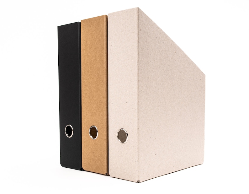 May include: Three file folders, one black, one brown, and one beige, are stacked on a white surface. The folders are made of cardboard and have metal rings for holding papers.